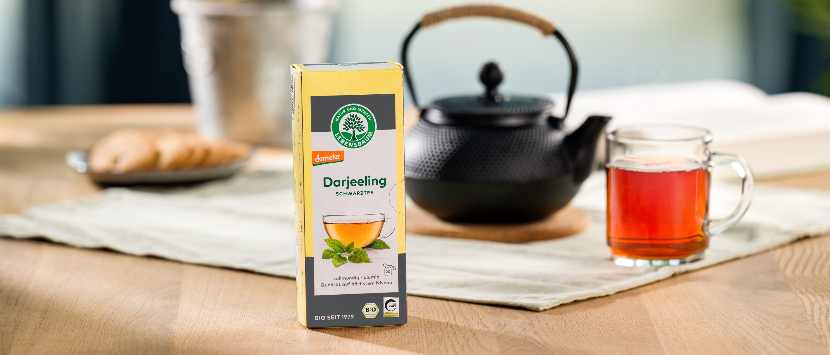 Eine Packung Lebensbaum Darjeeling liegt im Vordergrund, dahinter eine schwarze Teekanne und ein Glas mit frisch aufgebrühtem Tee auf einem hellen Tischläufer