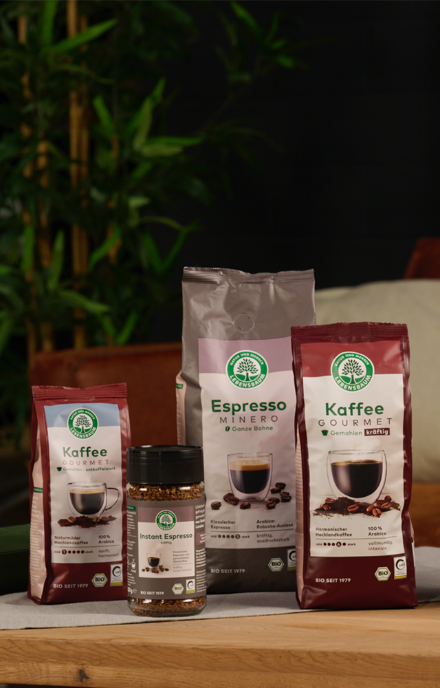 Verschiedene Kaffeeprodukte von Lebensbaum sind auf einem Holztisch arrangiert, im Hintergrund ein braunes Sofa und grüne Pflanzen