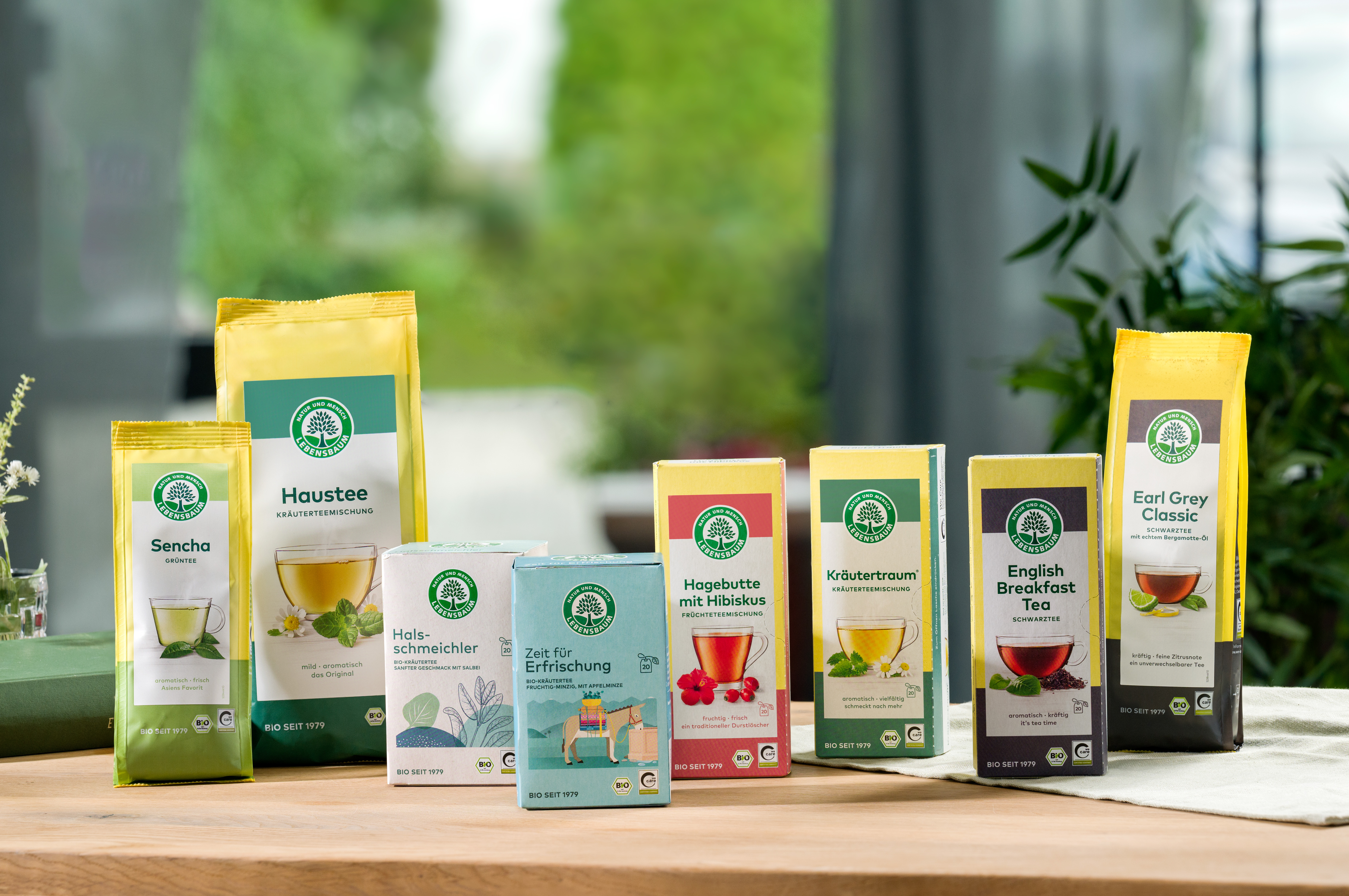 Teesortiment der Marke Lebensbaum mit Bio-Zertifizierung: Sencha, Haustee, Halsschmeichler, Zeit für Erfrischung, Hagebutte mit Hibiskus, Kräutertraum und Earl Grey Classic in gelben und farbigen Verpackungen auf Holztisch vor grünem Hintergrund.