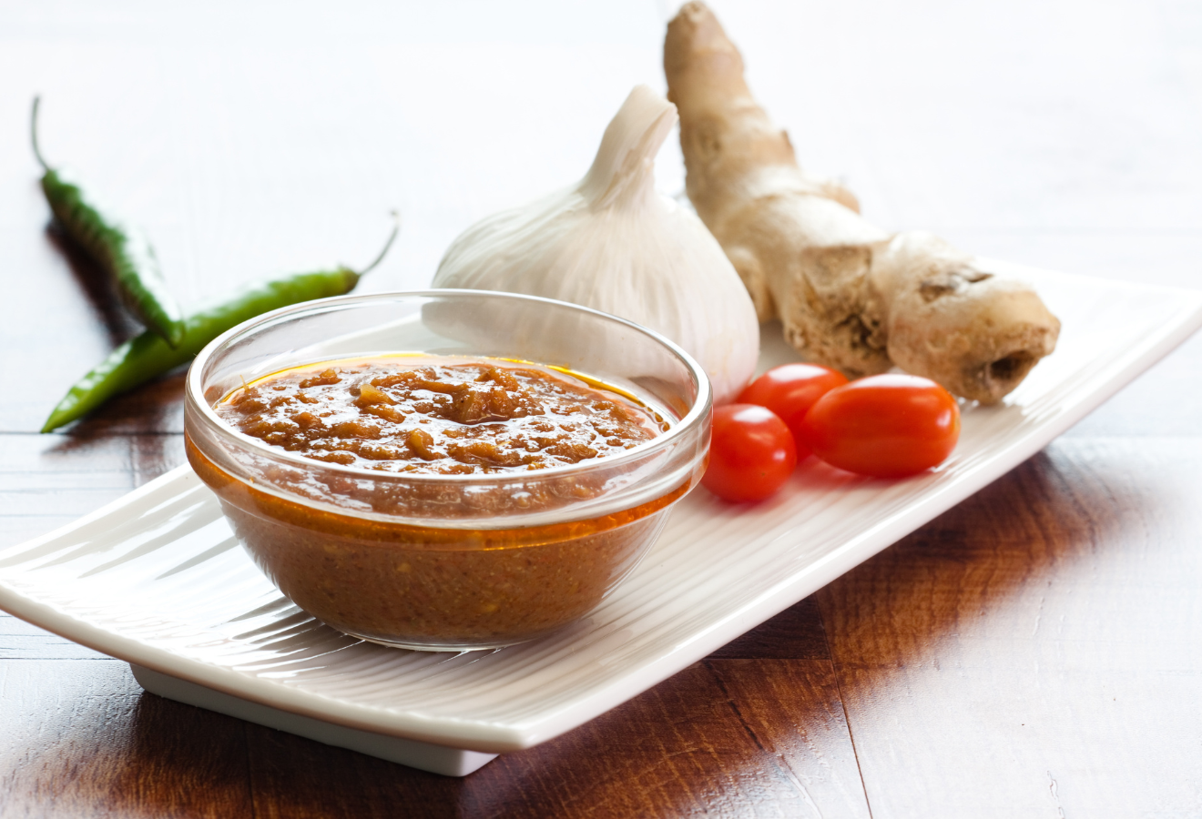 Kräuter-Marinade in einem Glasschälchen, dahinter Knoblauch, Ingwer und Tomaten