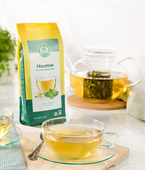 Packung Lebensbaum Haustee steht neben einer Teekanne mit aufgebrühtem Kräutertee und zwei Glastassen auf einem hellen Tisch