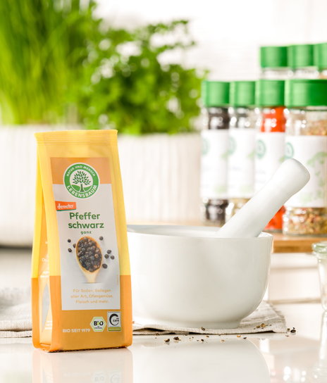 Packung Lebensbaum Pfeffer schwarz steht neben einem weißen Mörser auf einer hellen Küchenarbeitsfläche, im Hintergrund frische Kräuter und Gewürze