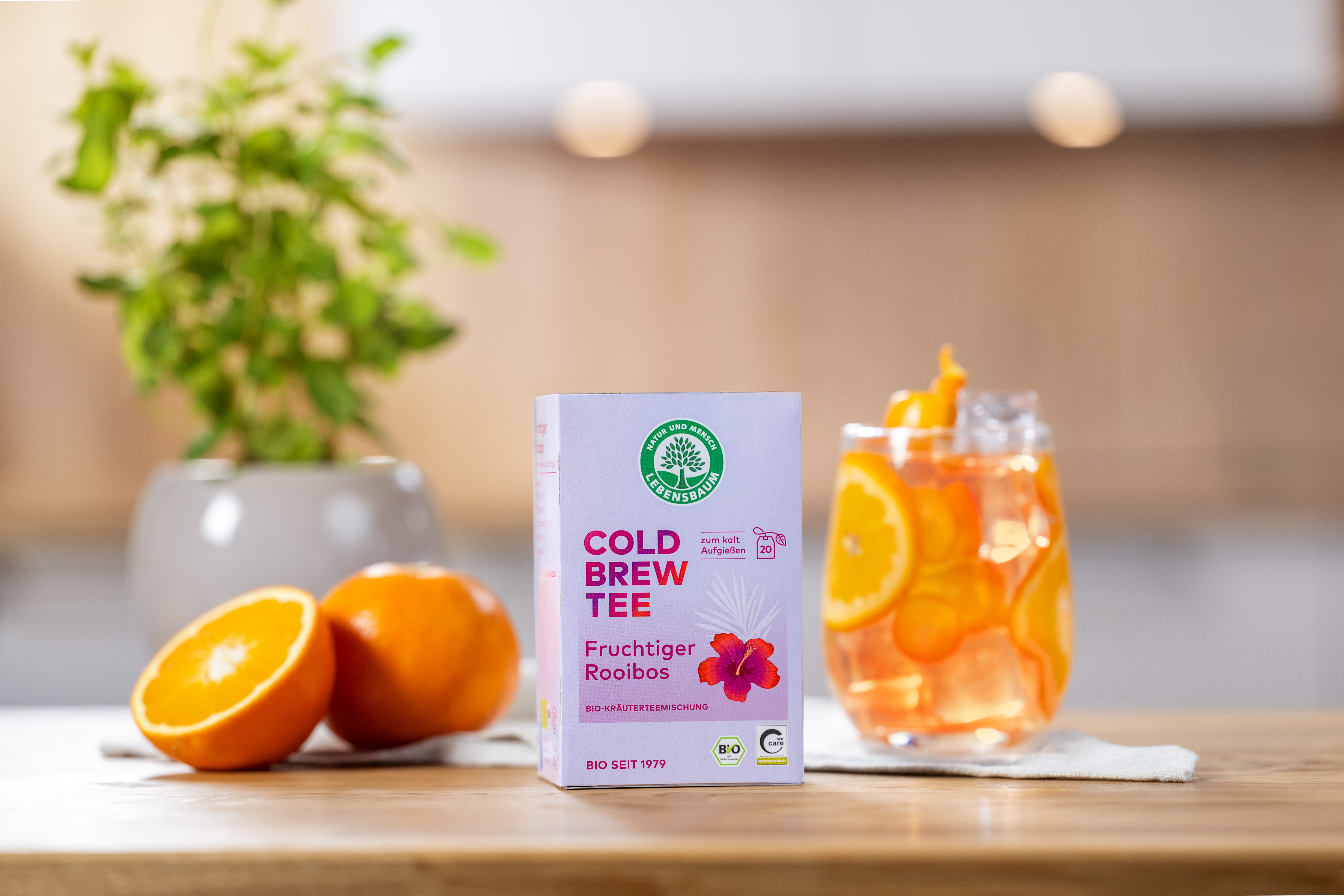 Bio-Cold-Brew-Tee fruchtiger Rooibos auf einem Tisch neben einem lecker aussehenden Glas n mit Cold-Brew und Eis, im Hintergrund eine Küche