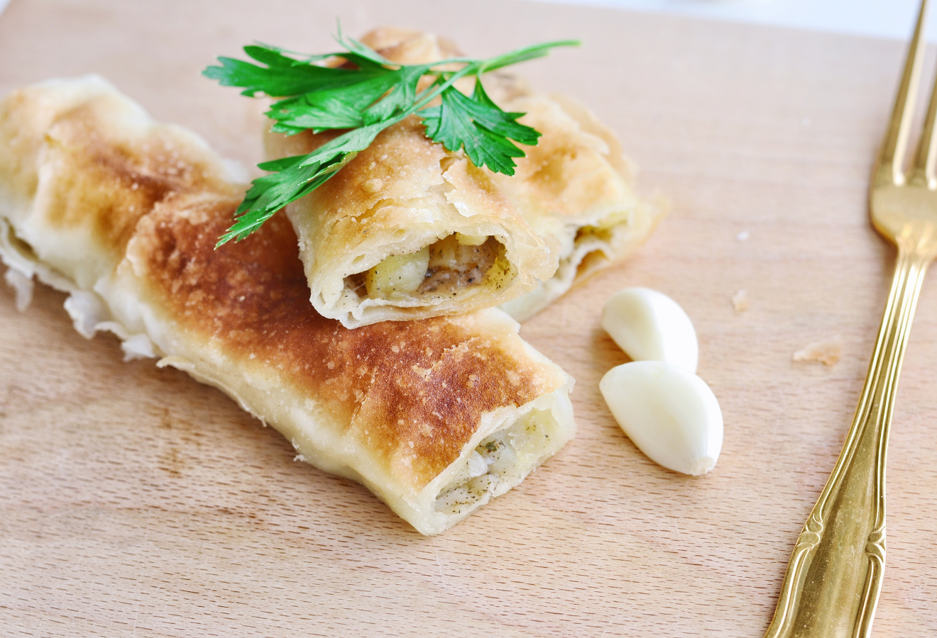 Vegetarischer Gemüsestrudel auf einem Holzbrett, davor zwei Knoblauchzehen und eine Gabel