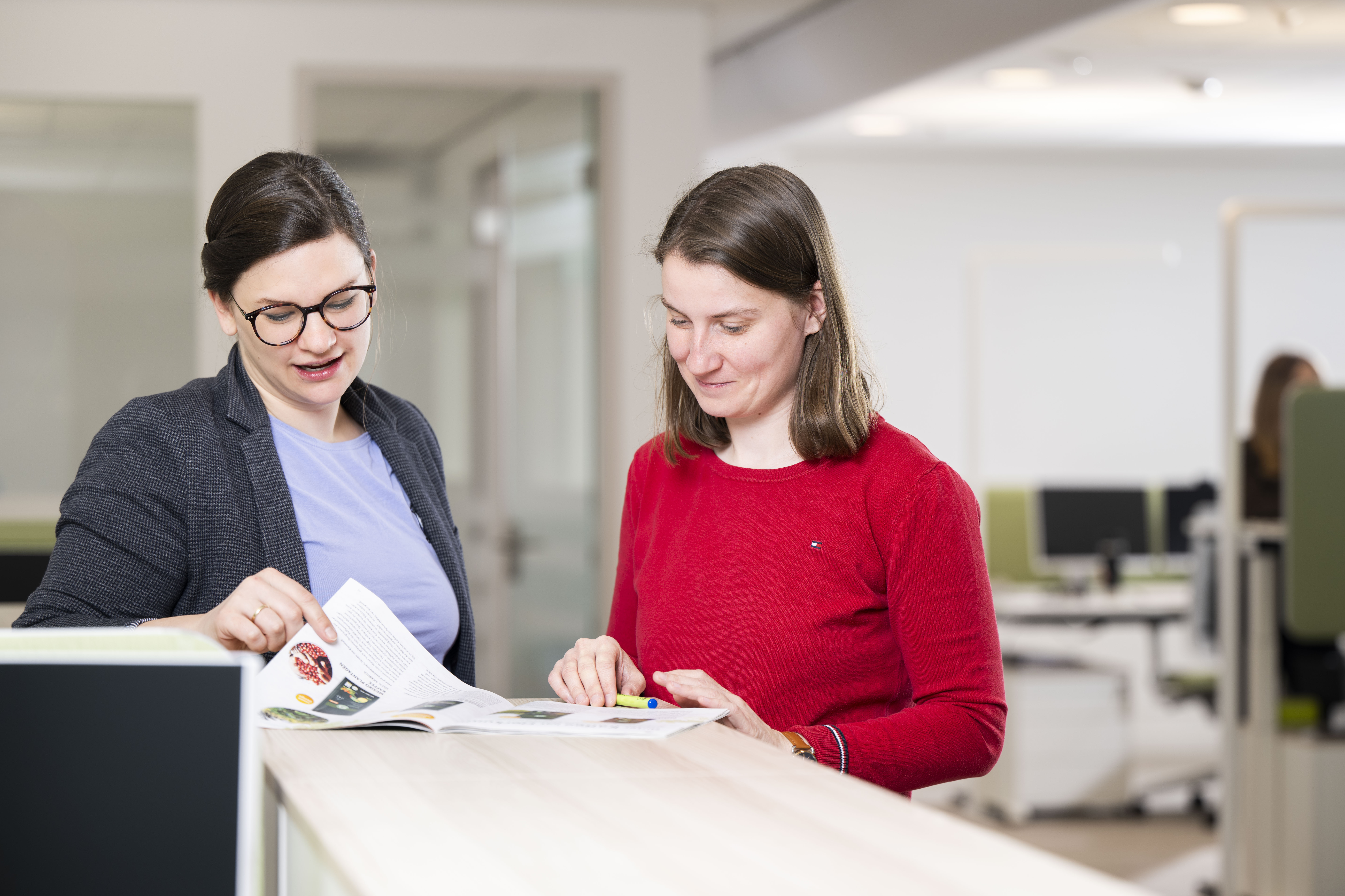 Zwei Mitarbeiterinnen stehen in einem Großraumbüro und betrachten gemeinsam eine aufgeschlagene Zeitschrift.
