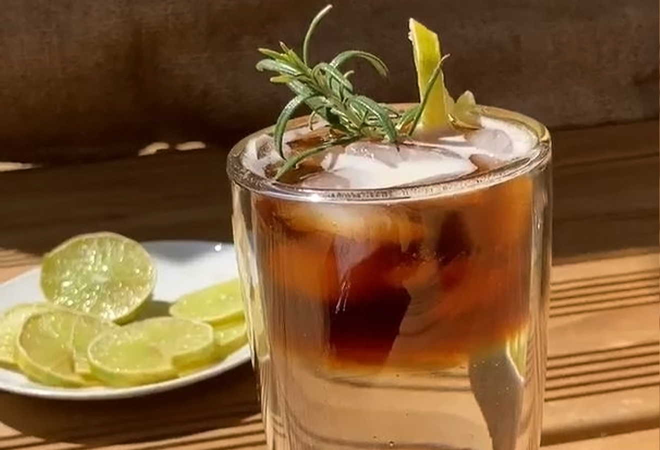 Espresso Tonic mit Eiswürfeln und Rosmarin in einem Glas, im Hintergrund Limettenscheiben auf einem Teller