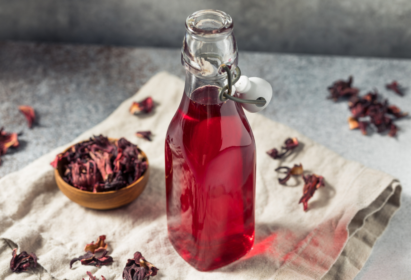 Sirup aus Hibiskusblüten in einer Glasflasche, dahinter ein Holzschälchen mit getrockneten Hibiskusblüten