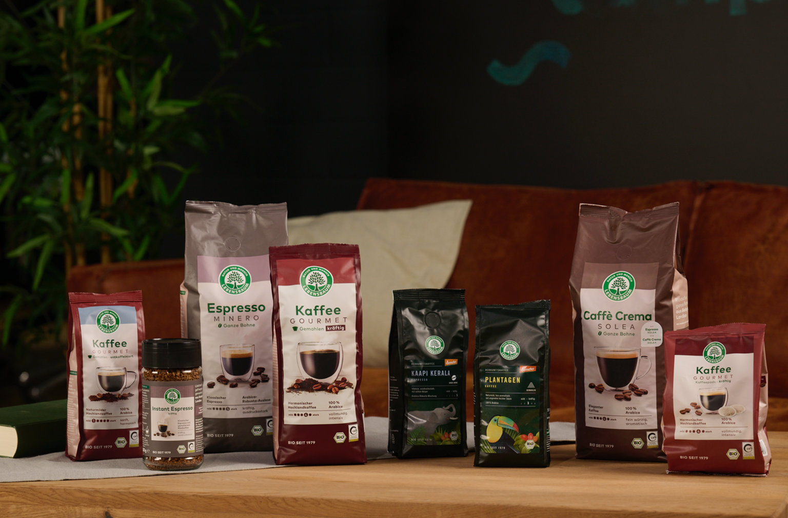Verschiedene Kaffeeprodukte von Lebensbaum sind auf einem Holztisch arrangiert, im Hintergrund ein braunes Sofa und grüne Pflanzen