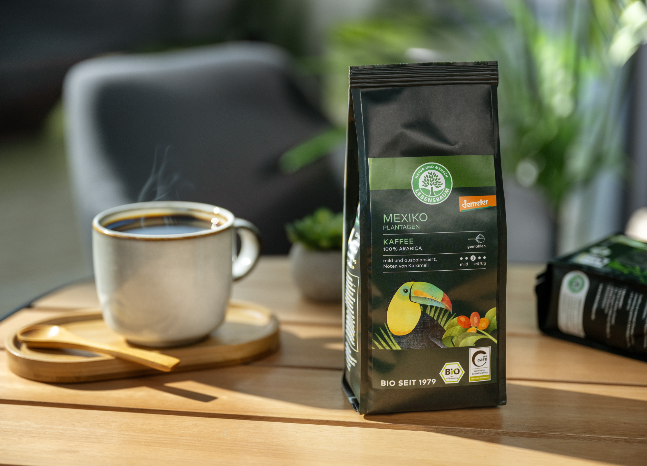 Eine Tasse dampfender Kaffee steht neben einer Packung Lebensbaum-Kaffee aus Mexiko auf einem Holztisch in sonniger Atmosphäre