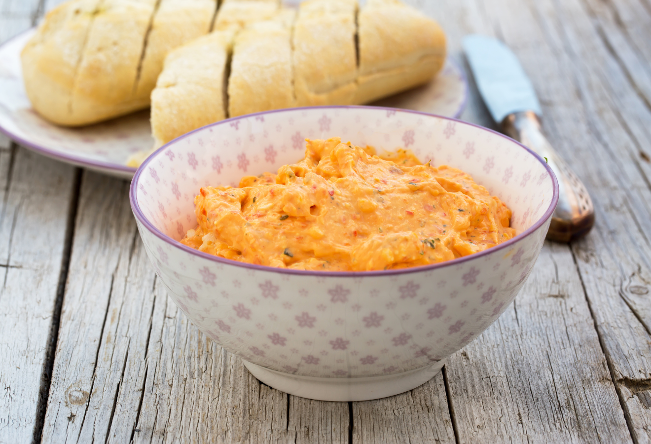 Rauchiger Paprika-Feta-Dip in einer weißen Schüssel, dahinter ein Teller mit Brot