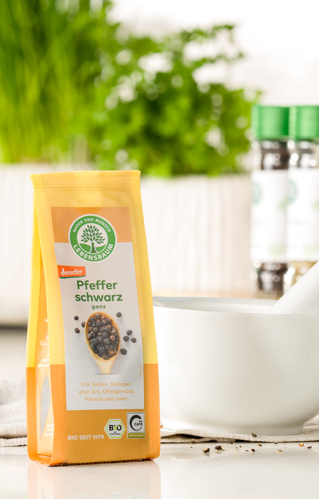 Packung Lebensbaum Pfeffer schwarz steht neben einem weißen Mörser auf einer hellen Küchenarbeitsfläche, im Hintergrund frische Kräuter und Gewürze