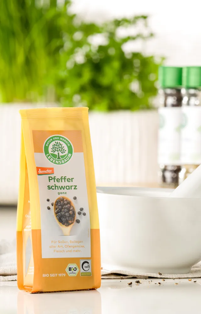 Packung Lebensbaum Pfeffer schwarz steht neben einem weißen Mörser auf einer hellen Küchenarbeitsfläche, im Hintergrund frische Kräuter und Gewürze