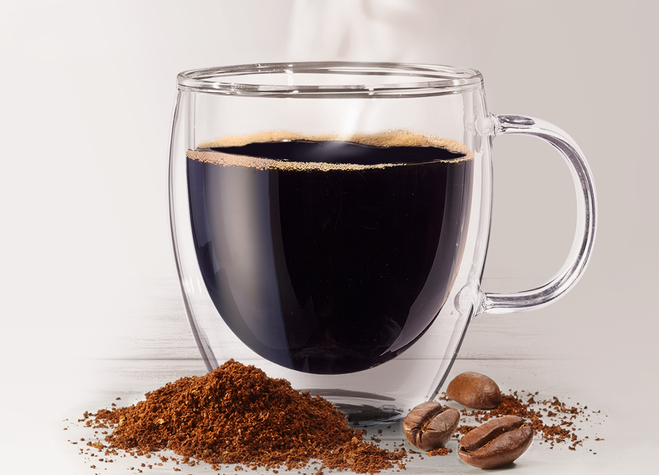 Ein doppelwandiges Glas mit schwarzem Kaffee, davor liegen Kaffeepulver und einige Kaffeebohnen auf einer hellen Oberfläche