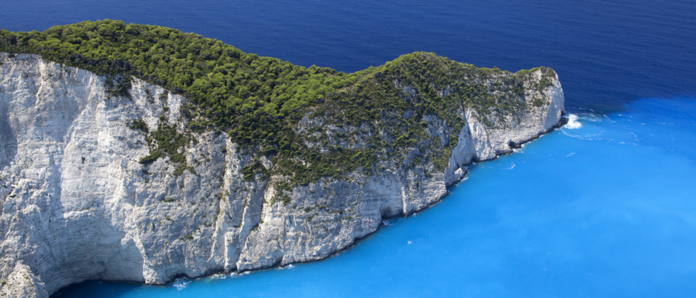 Weiße Steilküste mit dichtem Wald, umgeben von tiefblauem und türkisfarbenem Meer in Griechenland