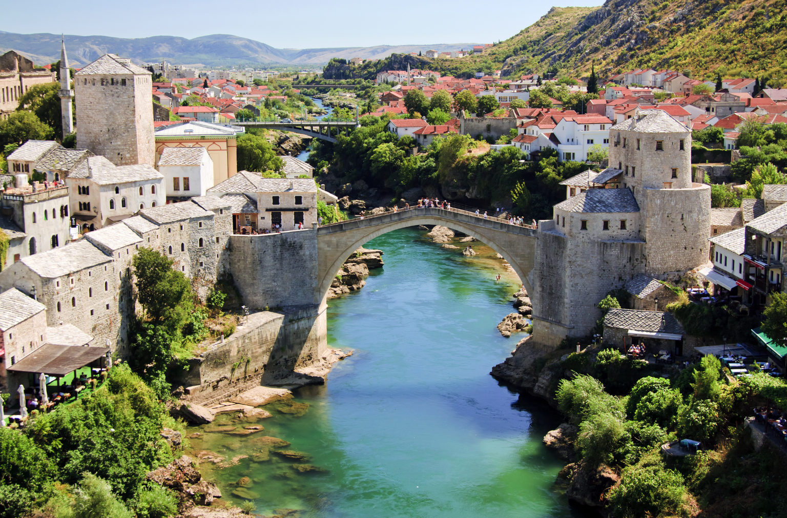 Historische Steinbrücke über einen Fluss inmitten einer Altstadt mit Hügeln im Hintergrund aus Bosnien Herzegowina