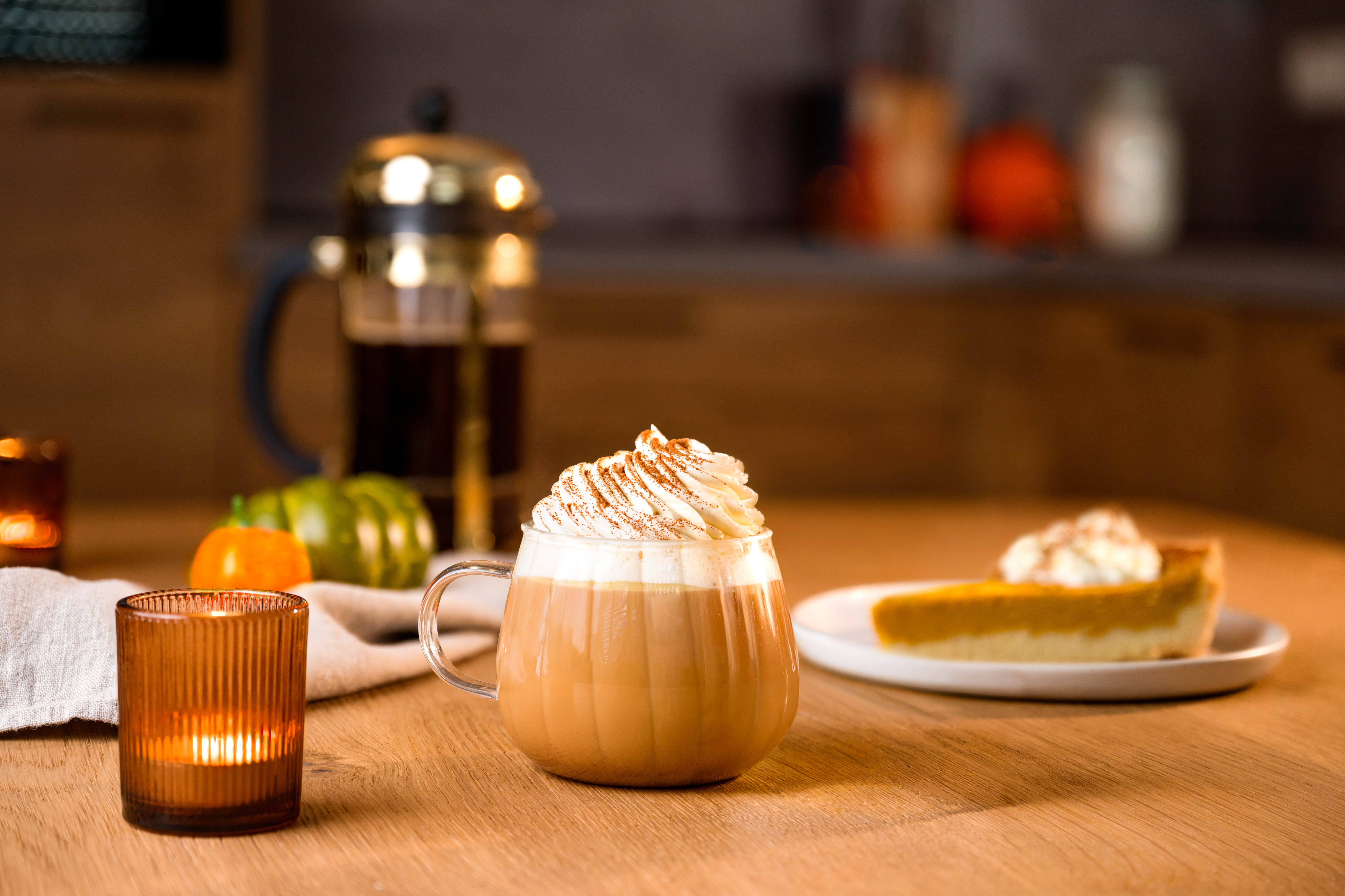 ein Glass Pumpkin Spice Latte mit Sahne steht auf einem Holztisch. Im Hintergrund sind eine Kaffeekanne und ein stück Kürbiskuchen