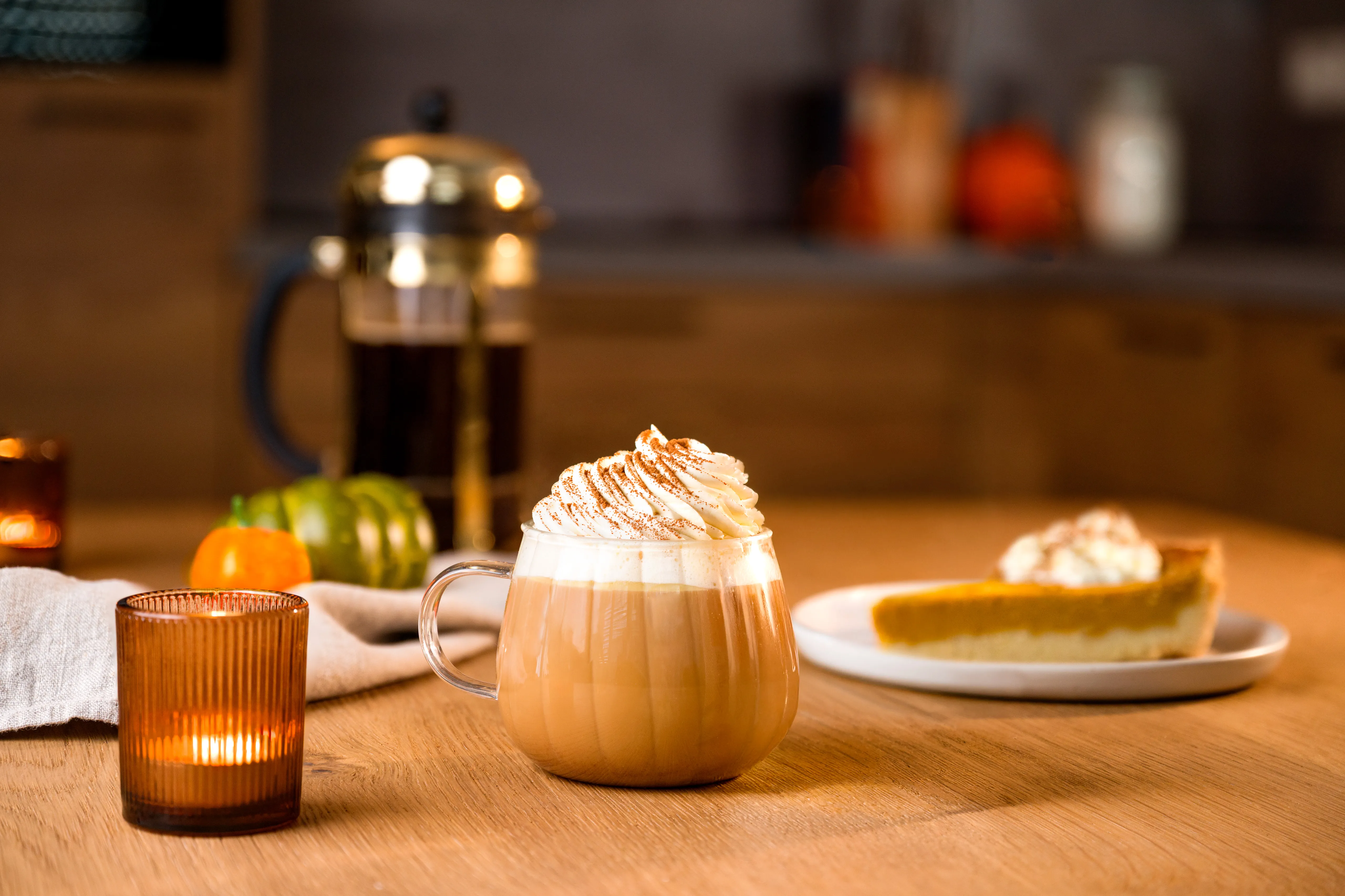 ein Glass Pumpkin Spice Latte mit Sahne steht auf einem Holztisch. Im Hintergrund sind eine Kaffeekanne und ein stück Kürbiskuchen