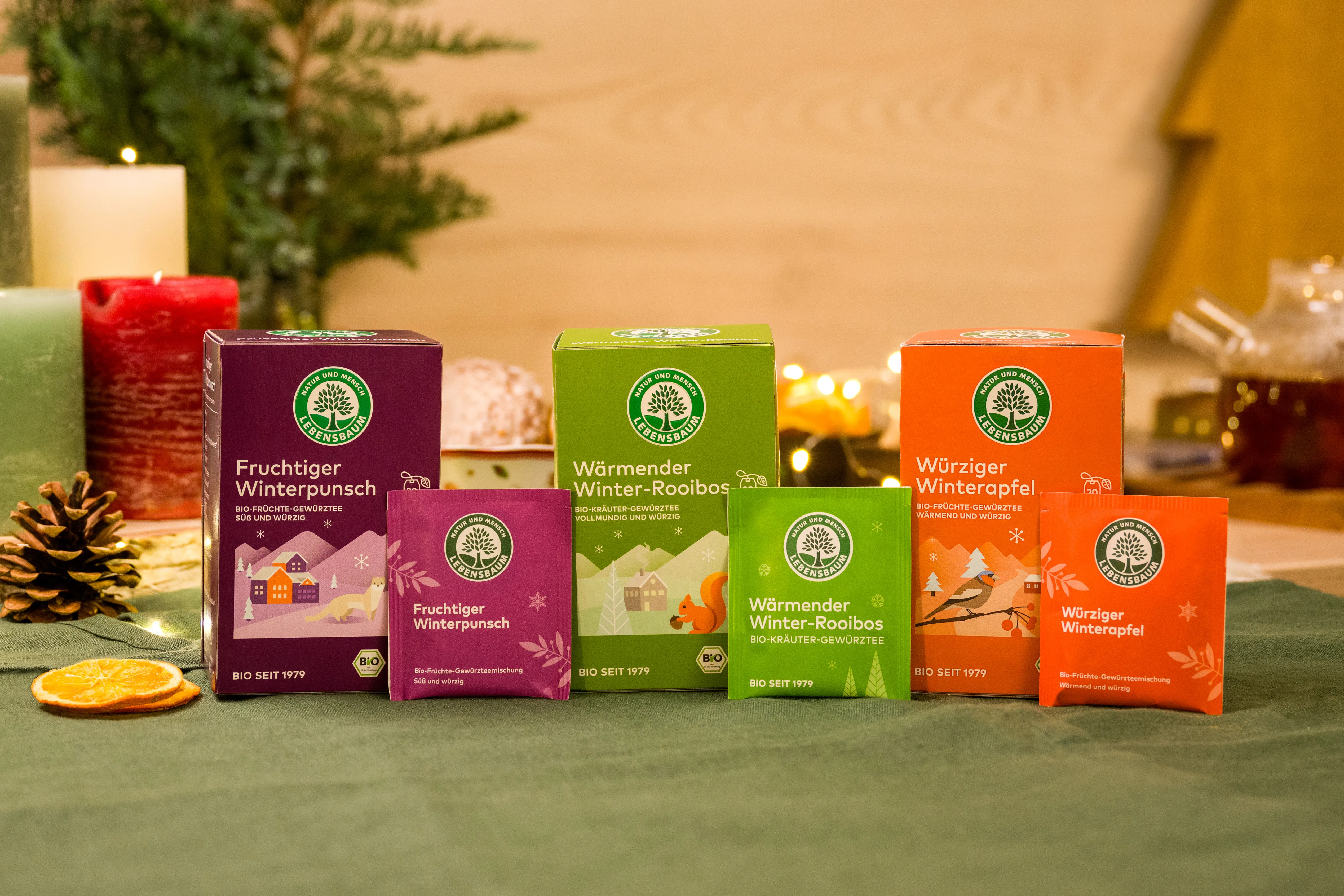 Bio-Tee-Kollektion in Winterverpackungen: Fruchtiger Winterpunsch (violett), Wärmender Winter-Rooibos (grün) und Würziger Winterapfel (orange) mit Dekoration.