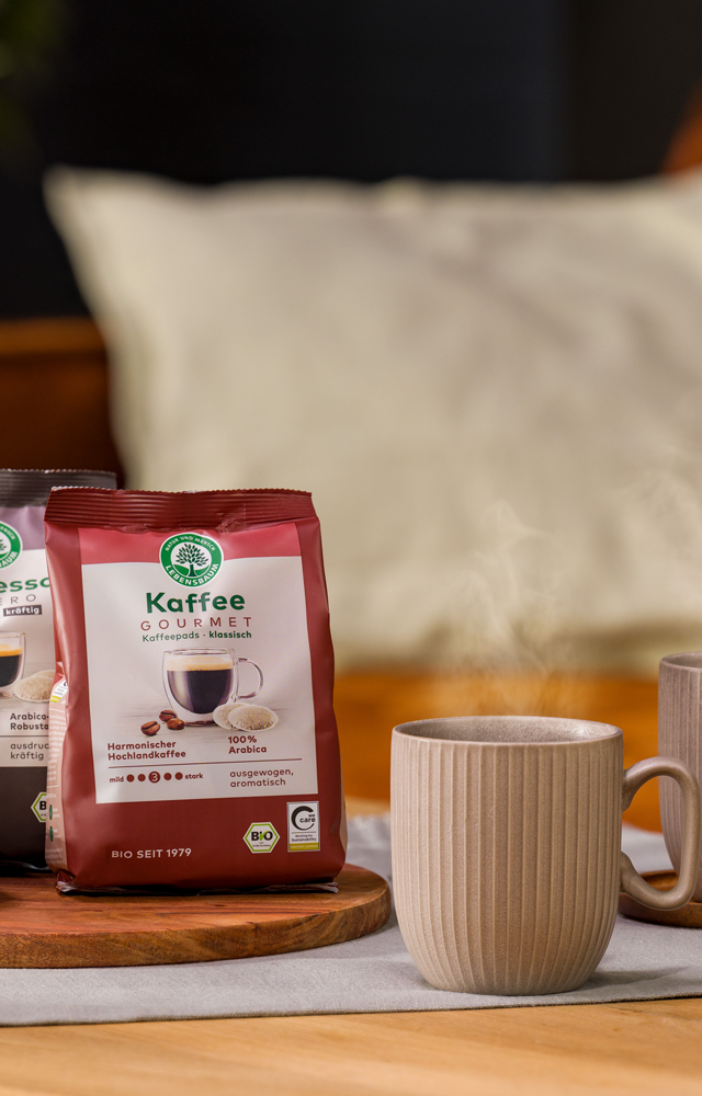 Zwei dampfende Tassen Kaffee stehen auf einem gedeckten Tisch neben einer Keks­schale und zwei Packungen Lebensbaum Kaffee und Espresso