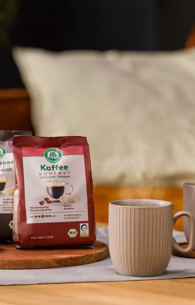 Zwei dampfende Tassen Kaffee stehen auf einem gedeckten Tisch neben einer Keks­schale und zwei Packungen Lebensbaum Kaffee und Espresso