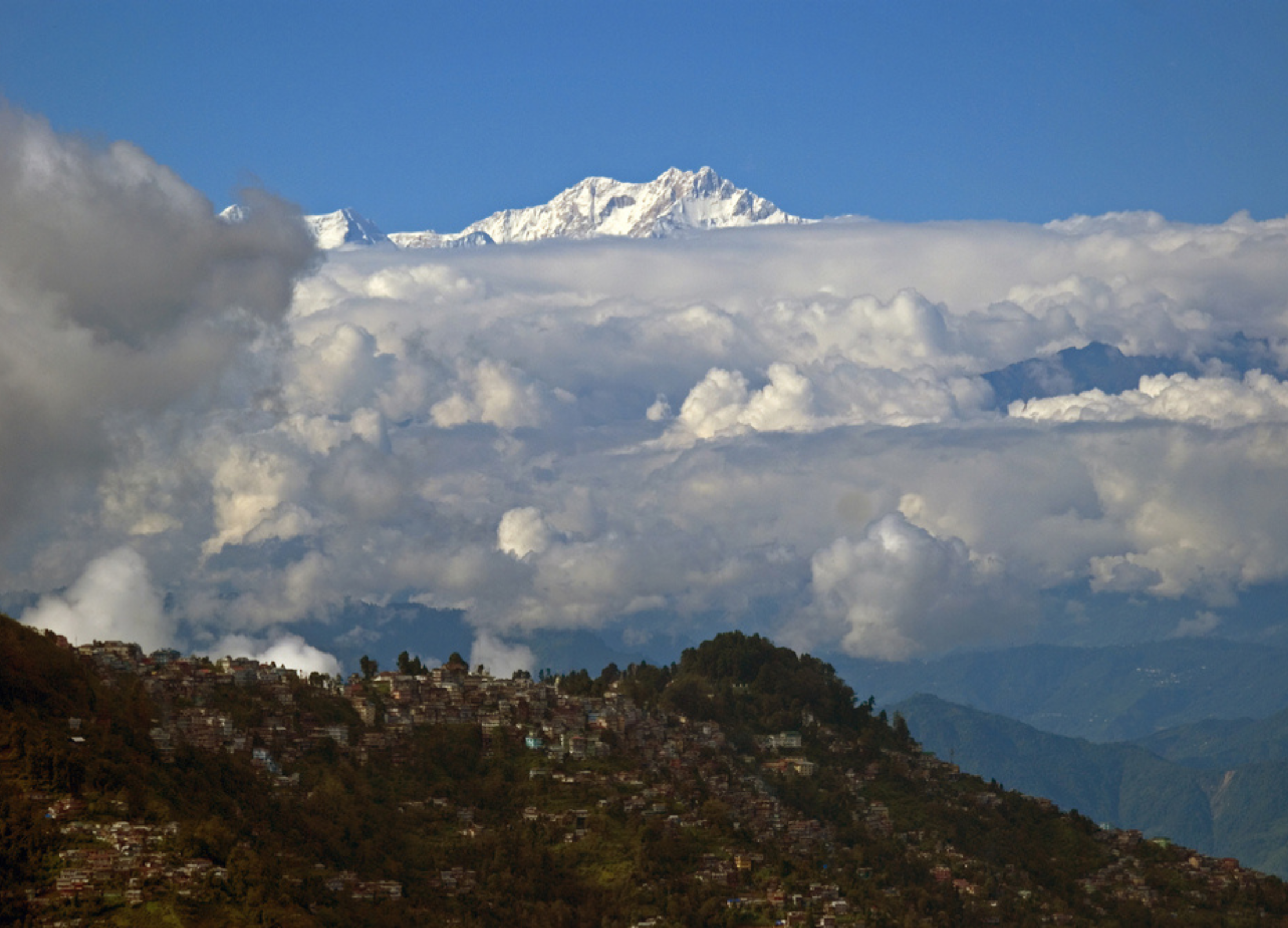 Schneebedeckter Berggipfel ragt über eine dichte Wolkendecke, darunter eine hügelige Stadtlandschaft