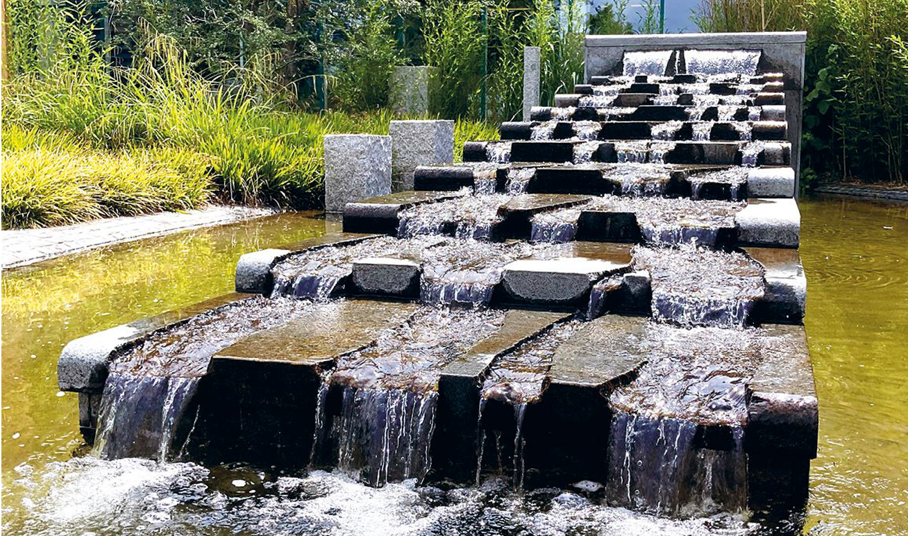 Stufenförmiger Wasserlauf aus Stein auf dem Gelände der Firma Lebensbaum, über den klares Wasser in ein flaches Becken plätschert, umgeben von grüner Bepflanzung