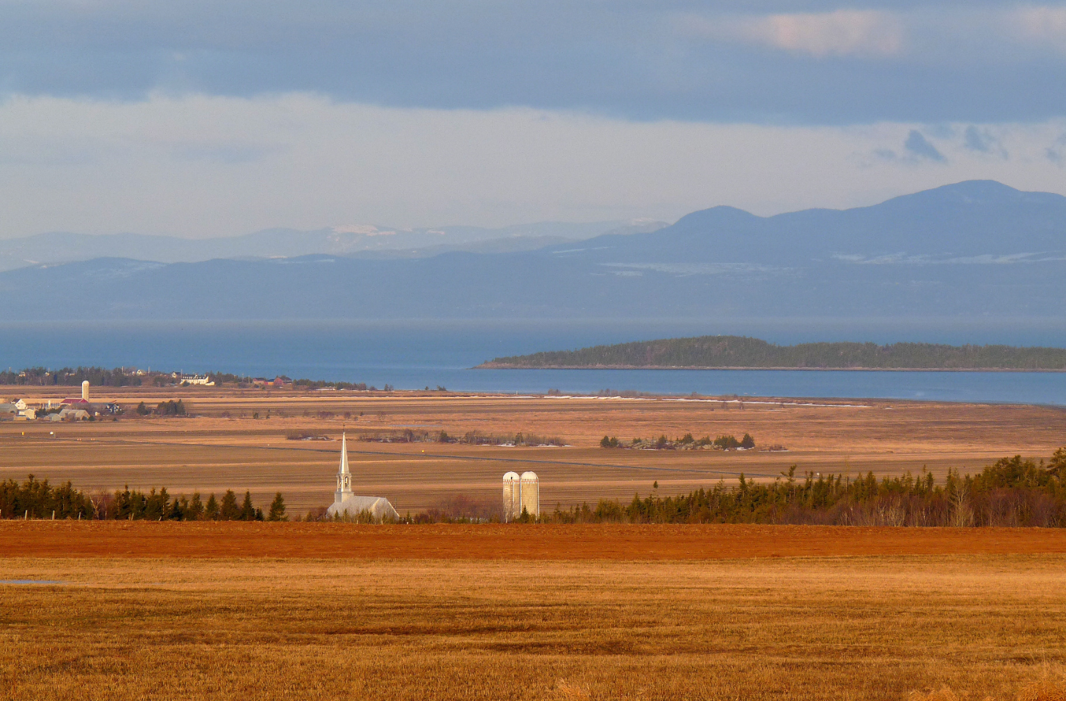 Weite Landschaft mit Feldern, Kirche, Wasser und Bergen im Hintergrund in Kanada