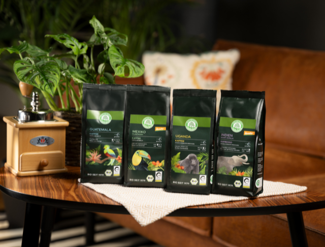 Herkunftskaffeepackungen mit Tiermotiven stehen auf einem Holztisch neben einer Kaffeemühle und einer Pflanze