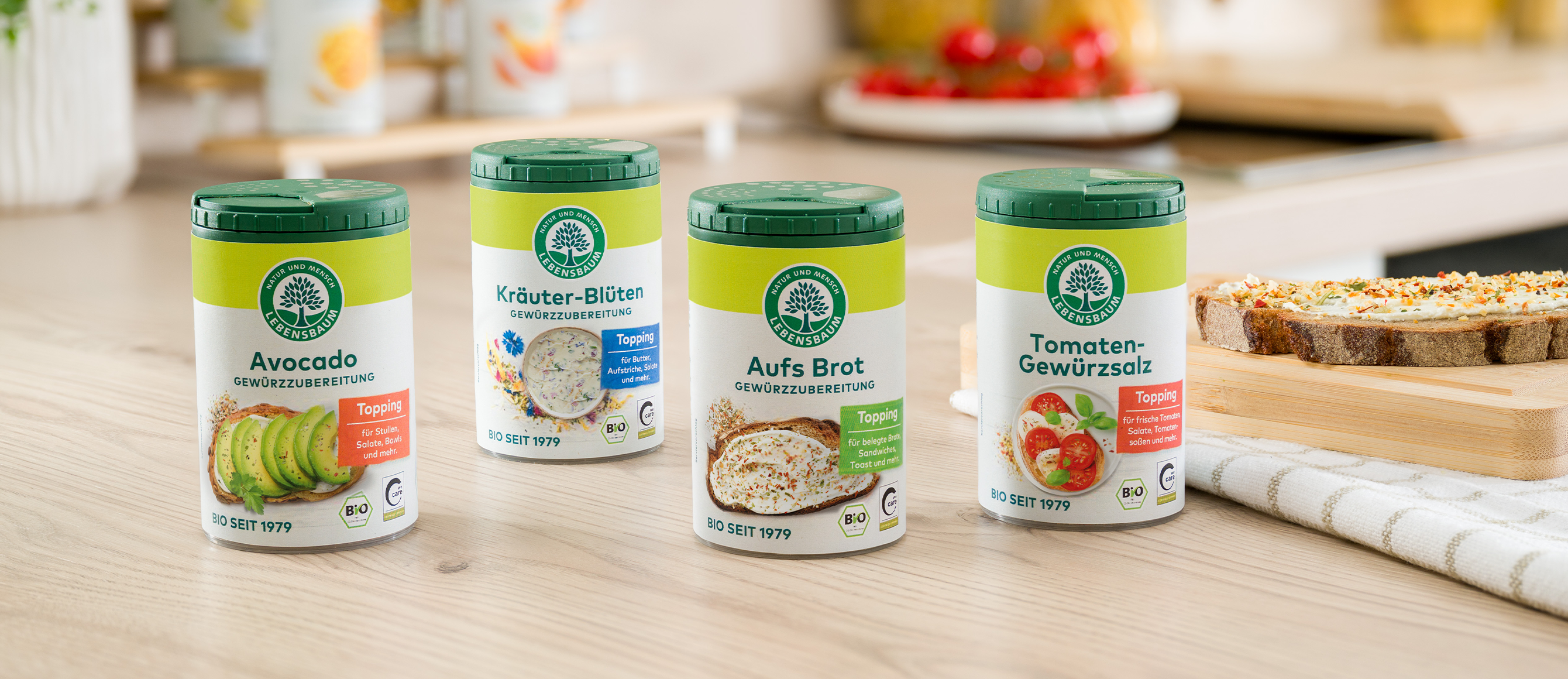 Vier Bio-Gewürztoppings der Marke Lebensbaum in grün-weißen Dosen: Avocado, Kräuter-Blüten, Aufs Brot und Tomaten-Gewürzsalz auf Holzarbeitsplatte mit Brotscheibe.